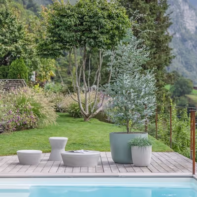 Swisspearl Kyoto Pflanzgefäße und Porto Tische sowie Hocker im Garten hinter einem Pool.